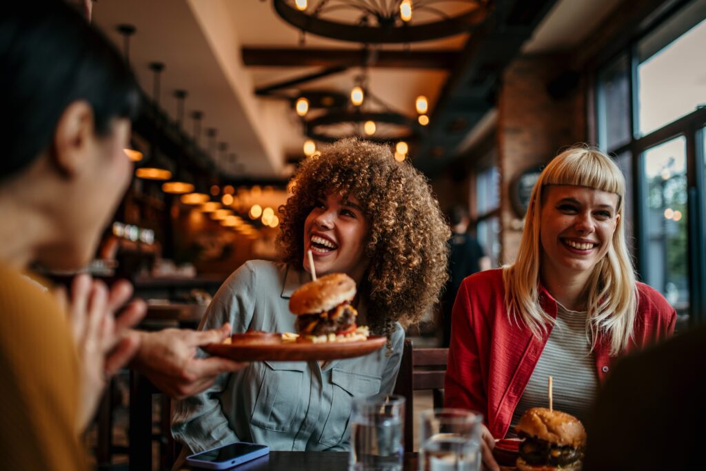 Three women sit at a restaurant table, smiling and laughing as a server delivers burgers. The setting is warmly lit with industrial-style décor and large windows. Glasses of water and phones are on the table.