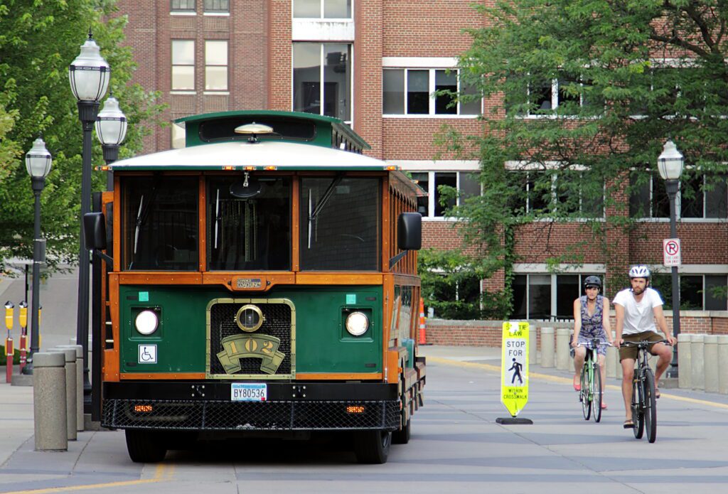 A green and orange trolley bus is stopped on a city street near cyclists. Two people ride bicycles past a sign that reads STOP FOR PEDESTRIANS in front of a brick building and trees.