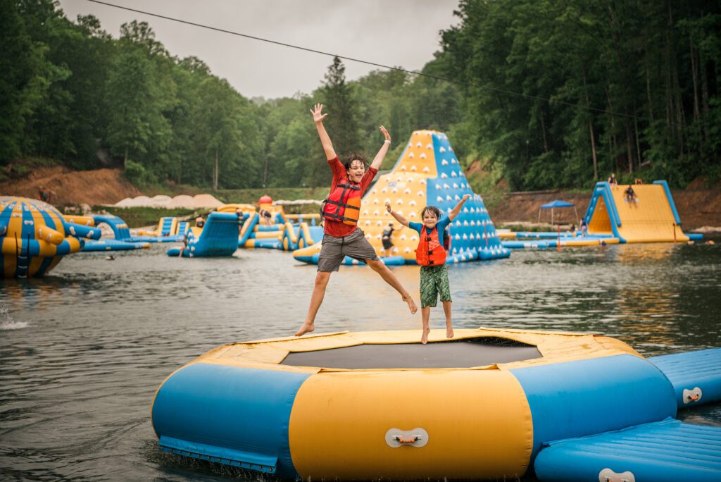 Two people wearing life jackets stand on a floating inflatable platform on a lake, with one jumping and the other smiling. Large inflatable structures and trees are visible in the background.