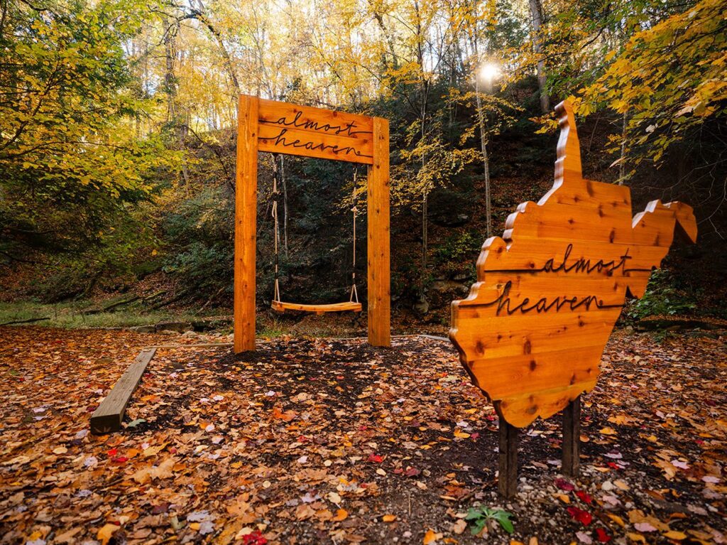 A wooden swing set in an autumn forest, with “almost heaven” written on the swing’s frame and a nearby wooden sign shaped like West Virginia, also inscribed with “almost heaven.” Sunlight filters through colorful fall leaves.