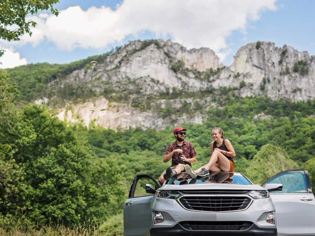 Two people sit on the hood of a silver SUV with open doors, smiling and talking, against a backdrop of green trees and a scenic, rocky mountain under a partly cloudy sky.