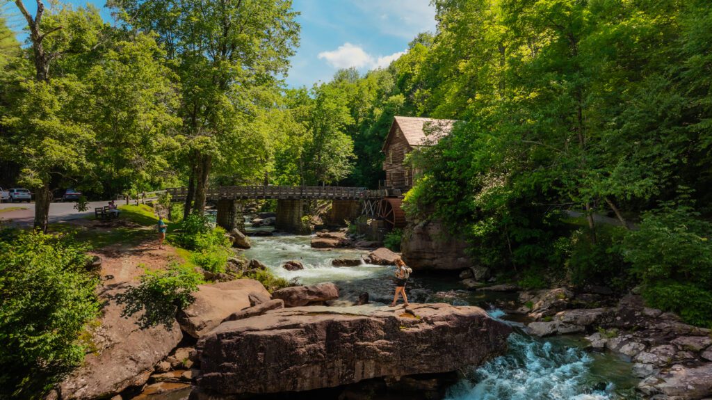 A person stands on a large rock by a rushing river, surrounded by lush green trees. A wooden bridge and an old mill house are in the background under a bright blue sky with scattered clouds.