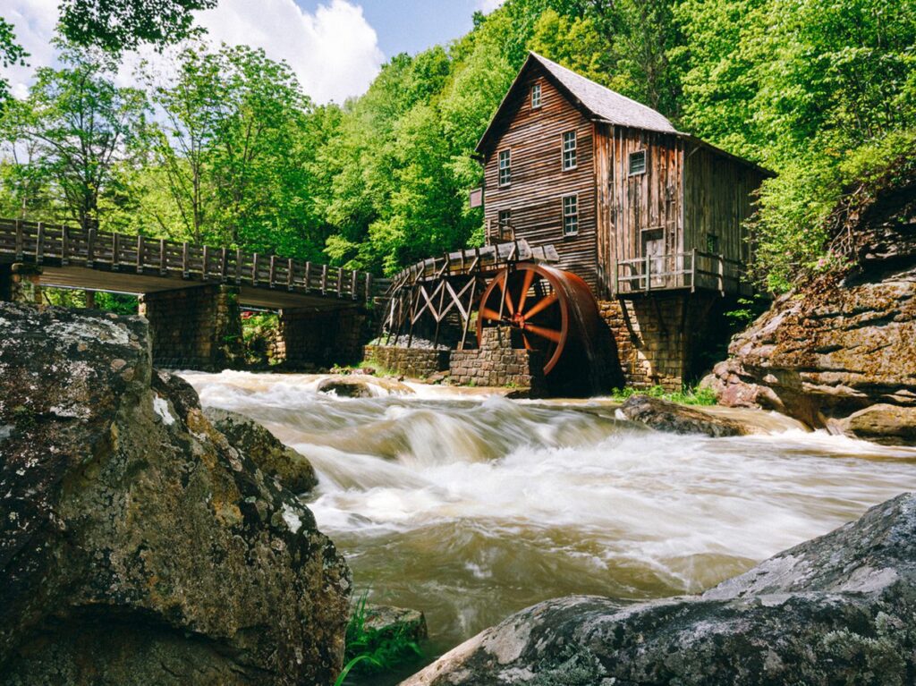 A rustic wooden watermill with a large red water wheel sits beside a fast-flowing river, surrounded by lush green trees and rocky shores under a partly cloudy sky.