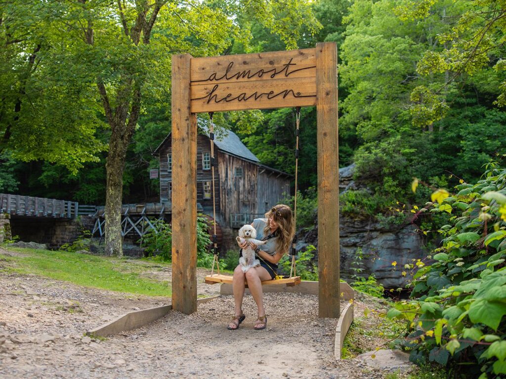 A woman sits on a wooden swing with a small white dog under a sign that reads “almost heaven.” Lush greenery and a rustic wooden mill are in the background.