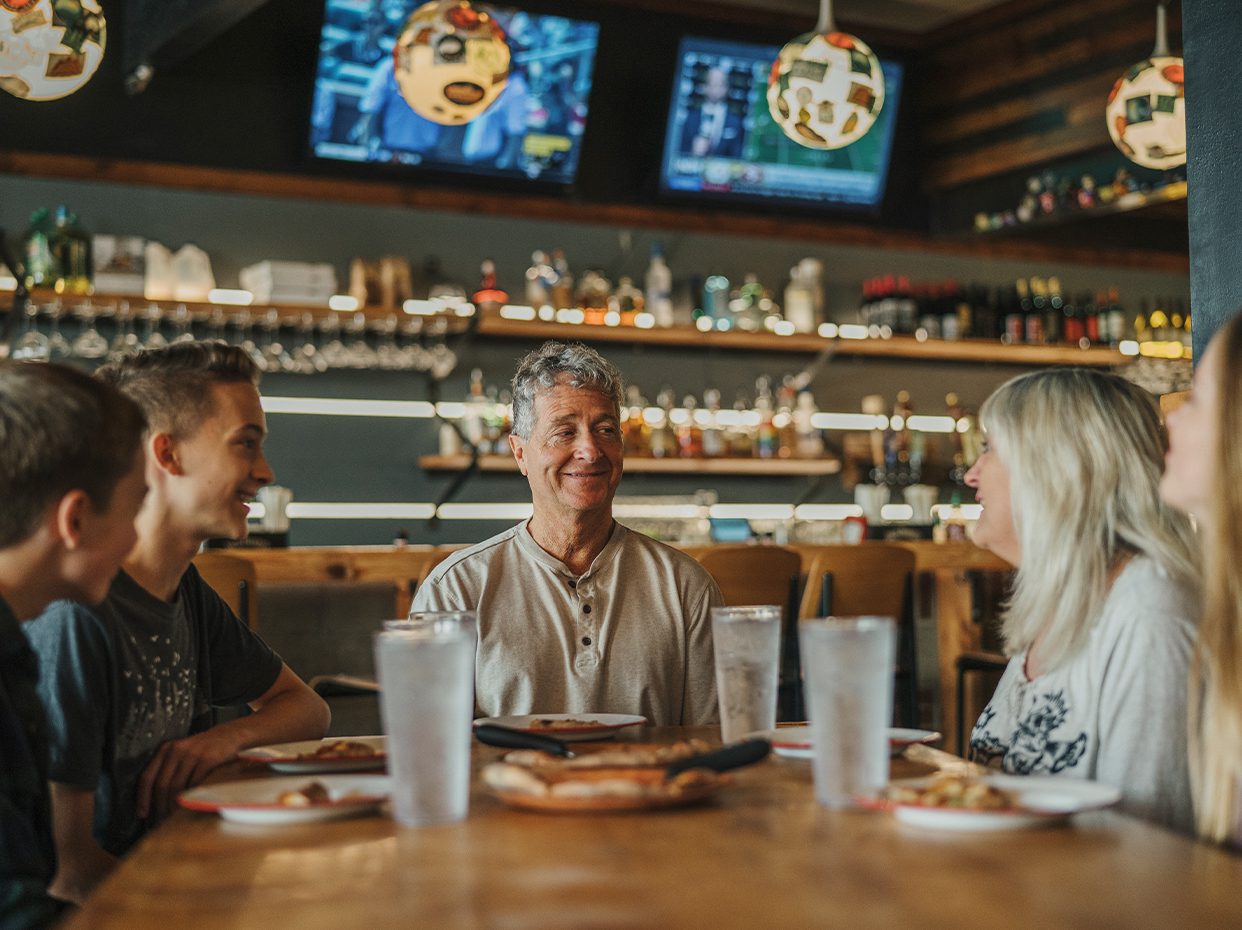 A group of people sit around a restaurant table, smiling and talking, with food and drinks in front of them. Shelves with bottles and two TVs are visible in the background.