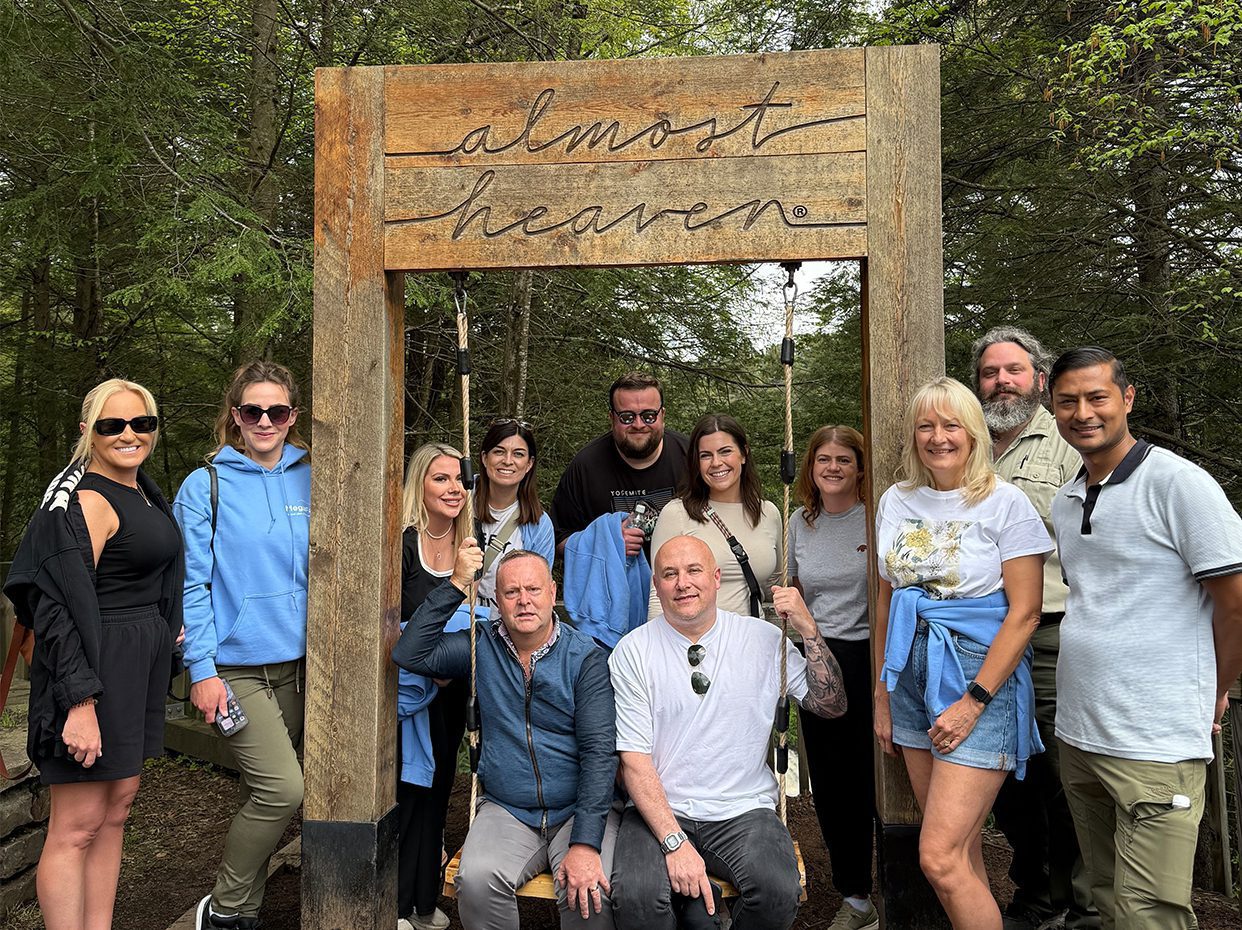 A group of thirteen people pose and smile together outdoors under a large wooden sign that reads almost heaven, surrounded by green trees and forest.