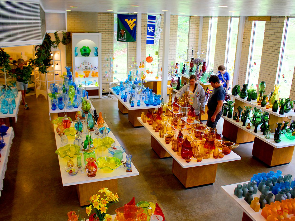 A bright glass shop interior displays colorful glass vases and art pieces on tables. Several people browse the items. Large windows let in natural light, and college flags hang on the back wall.