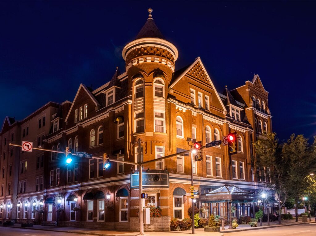 A historic, turreted red-brick building with peaked roofs and ornate trim is illuminated at night on a quiet street corner with traffic lights and street signs.