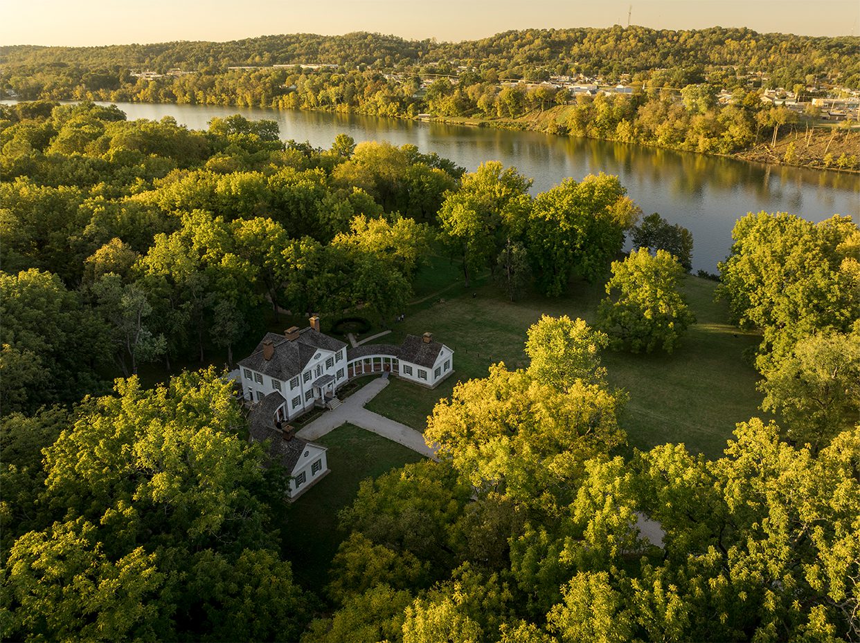 Aerial view of a historic white house surrounded by dense green trees near a wide river, with hills and a town visible in the distance under a golden evening light.
