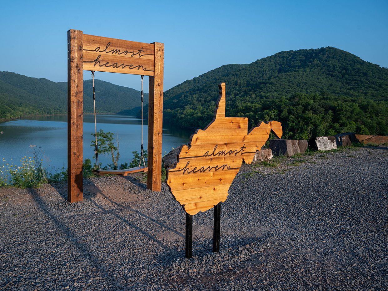 A wooden swing and a wooden cutout of West Virginia, both with almost heaven written on them, sit on a gravel area overlooking a lake and green, tree-covered hills under a clear blue sky.