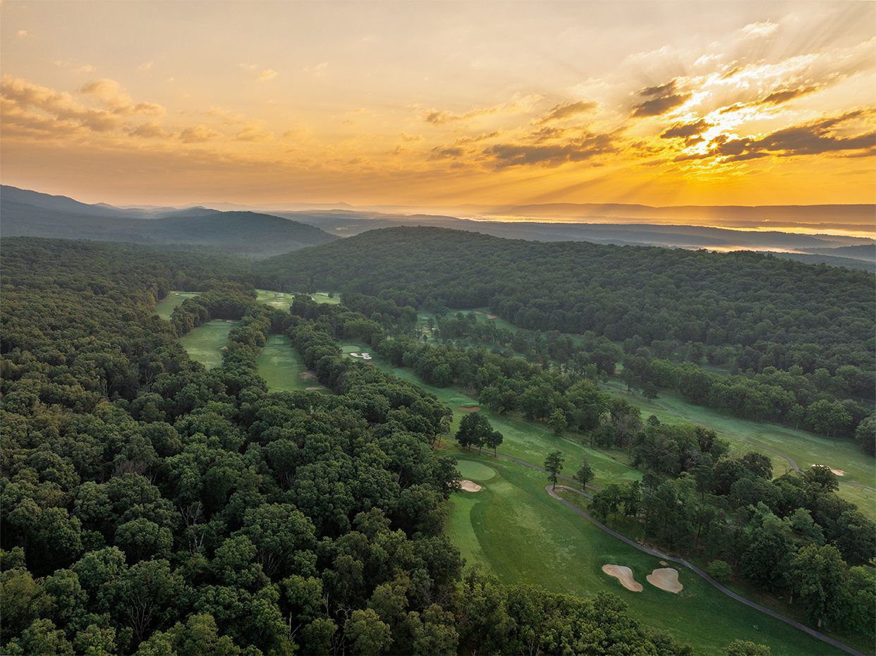 Aerial view of a lush, green golf course surrounded by dense forest, with mountains and a lake in the distance beneath a dramatic, golden sunset sky.