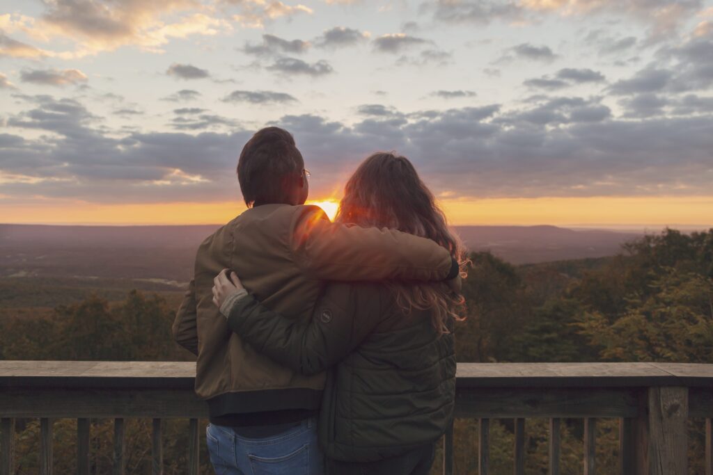 Two people stand on a wooden deck, hugging and looking at a scenic sunset over distant hills and trees with a partly cloudy sky.