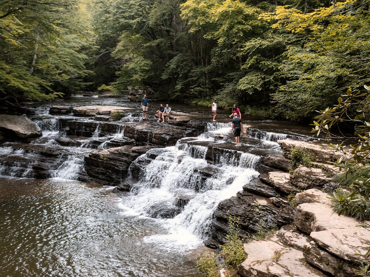 Several people stand and sit on layered rocks beside and above a small, gentle waterfall in a forested area with lush green trees surrounding a clear, shallow stream.