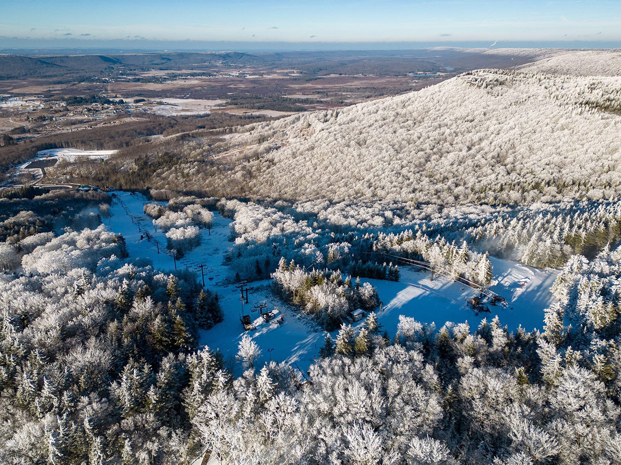 Aerial view of a snow-covered forested mountain, with ski slopes and ski lifts visible. The landscape extends into a valley with scattered trees and clearings under a bright blue sky.