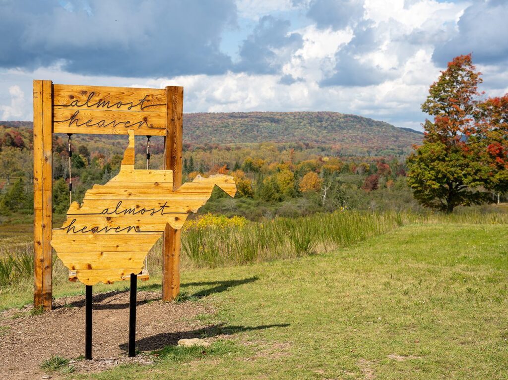 A wooden sign shaped like West Virginia with the words almost heaven stands in a grassy field. Colorful autumn trees and hills are visible in the background under a partly cloudy sky.