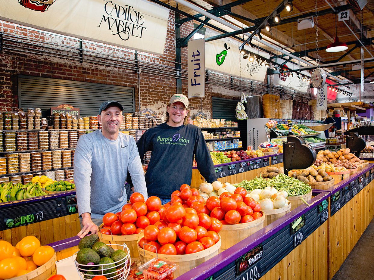 Two smiling vendors stand behind a colorful display of fresh produce, including tomatoes and peppers, at a well-lit indoor market labeled Capitol Market, with fruits and grocery items visible in the background.