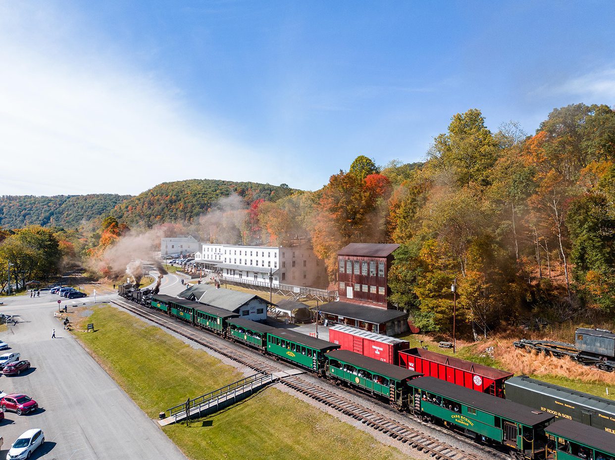 A scenic train with green passenger cars emits smoke as it passes through a small town surrounded by autumn trees and hills. Historic buildings and parked cars are visible near the train tracks.