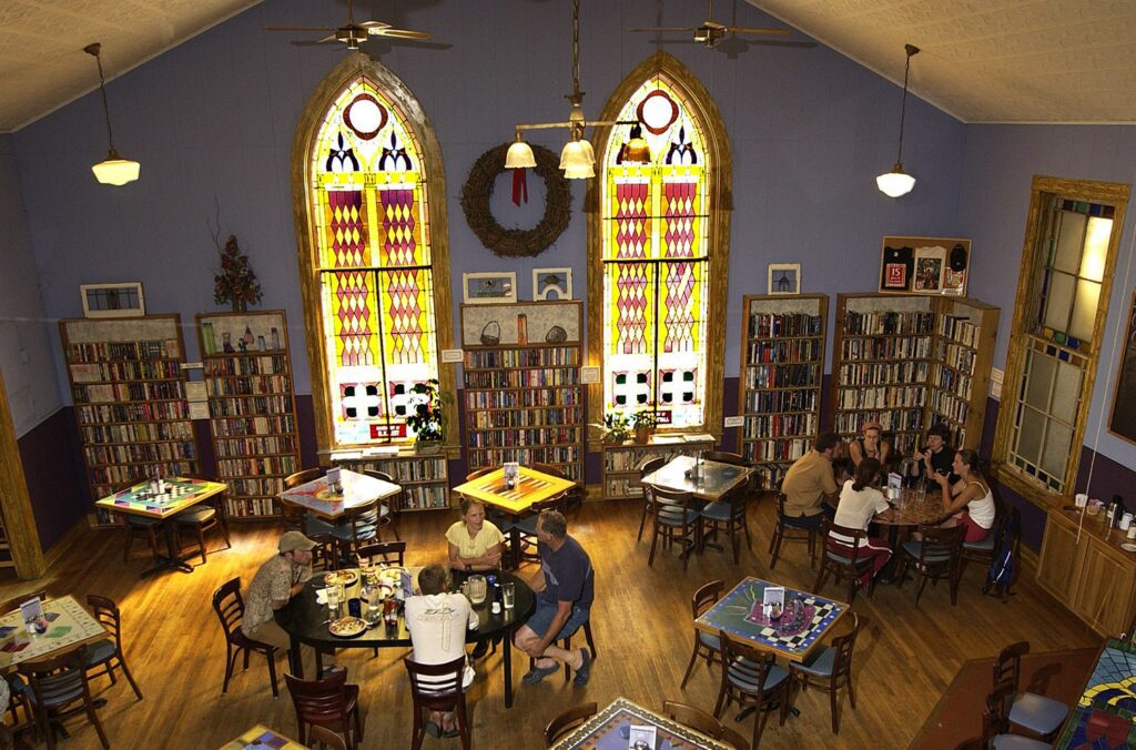 A cozy café with stained glass windows and bookshelves lining the walls. Several groups of people sit at wooden tables, eating and chatting, in a warmly lit, inviting atmosphere.
