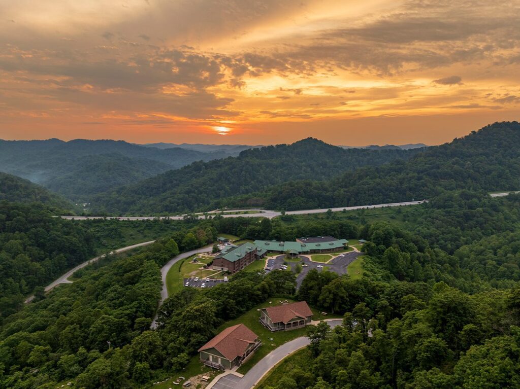 Aerial view of a large complex with multiple buildings surrounded by lush green hills and winding roads at sunset, with a dramatic sky and mountains in the distance.
