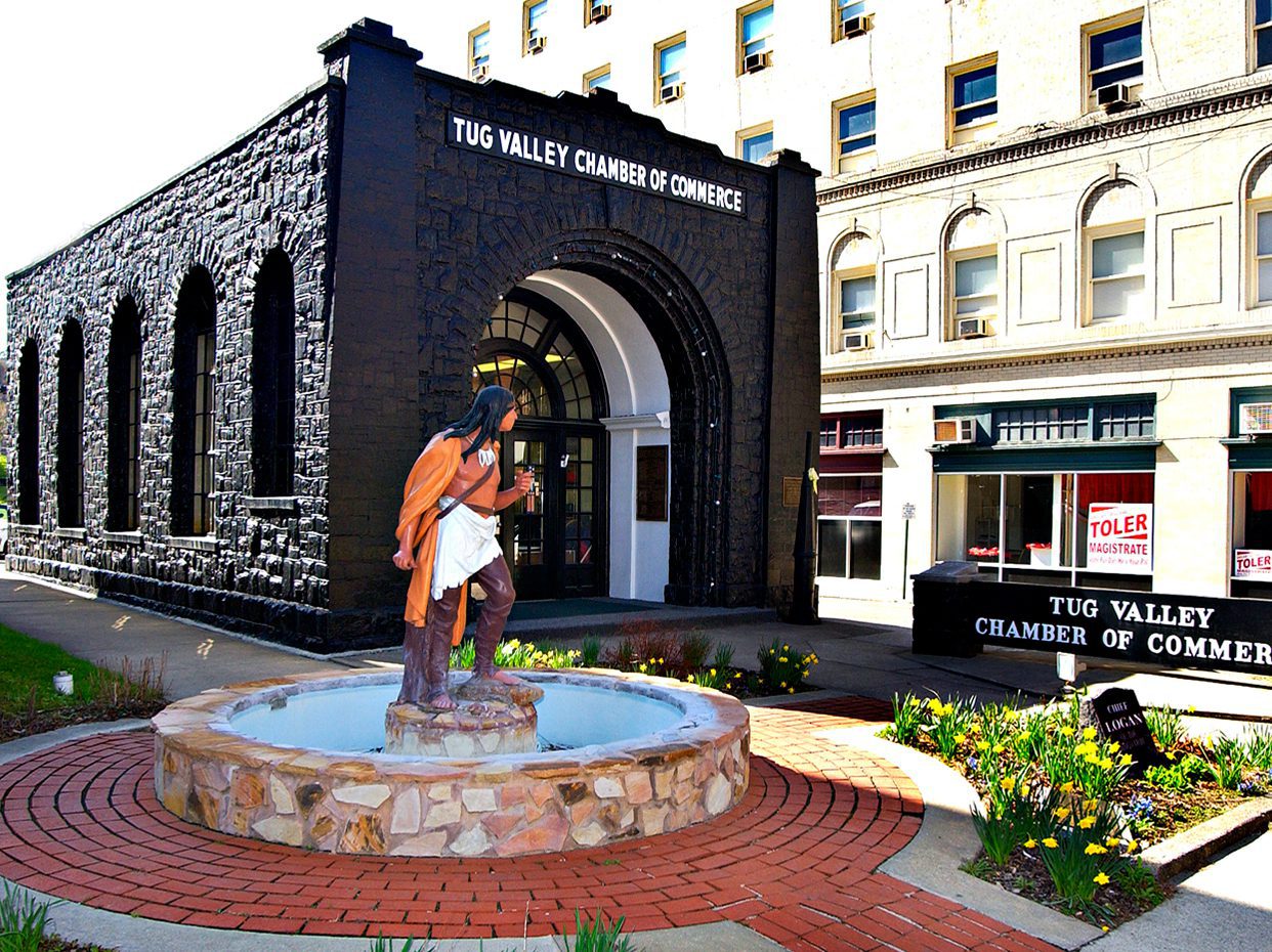 A statue of a Native American stands in a circular stone fountain in front of the Tug Valley Chamber of Commerce, housed in a black stone building next to a light-colored, multi-story structure.