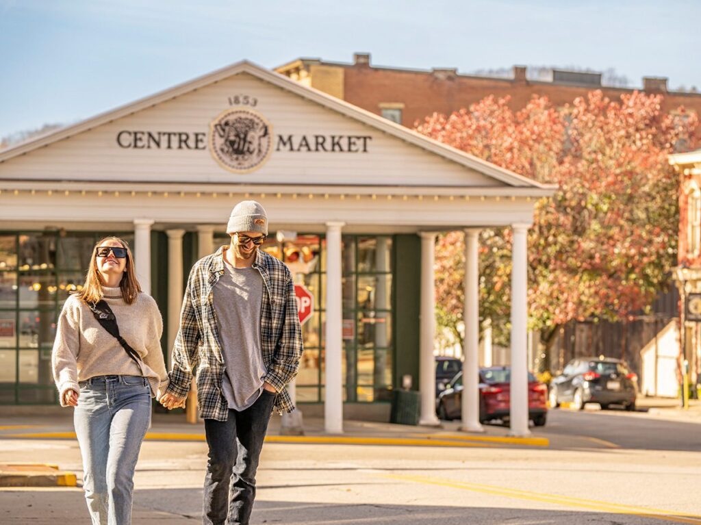 A couple holding hands and smiling walks outside Centre Market, a historic building with white columns and the year 1853 displayed above the entrance, on a sunny day with autumn trees in the background.