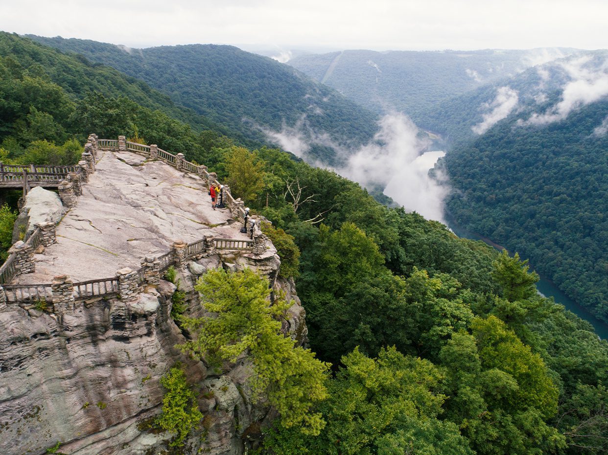 A rocky overlook with wooden railings juts out above a lush, green valley, with two people standing near the edge. Mist rises from the trees, and a river winds through the forested hills below.