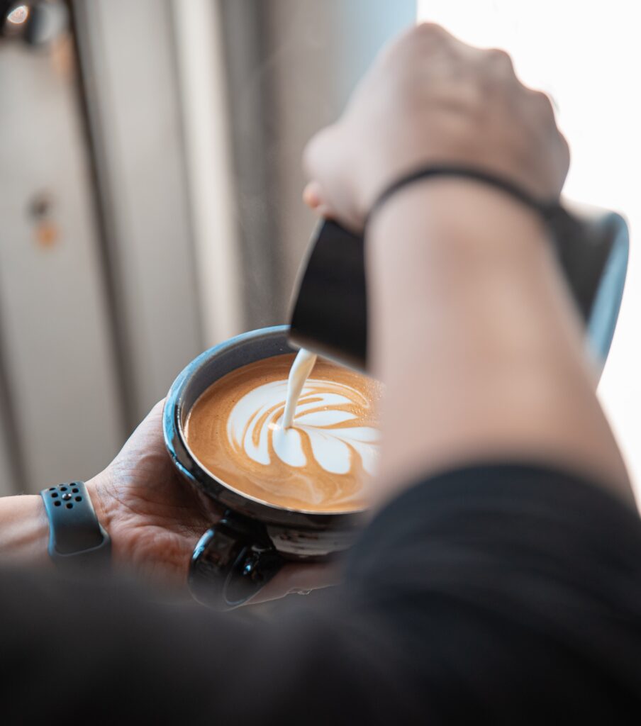 A person pours steamed milk into a cup of coffee, creating latte art in the shape of a leaf. The scene is viewed from above their shoulder, with the persons hands and blue wristband visible.
