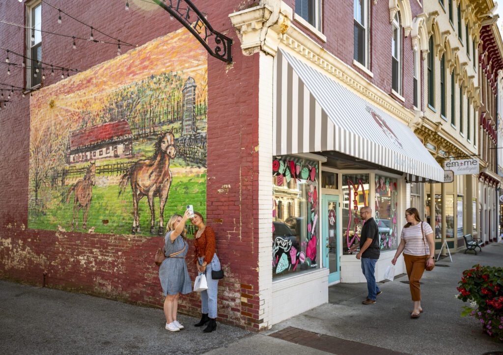 Two women pose taking a selfie in front of a colorful mural of horses on a brick building corner, while two other people stand by the entrance of a boutique with striped awning on a sunny day.