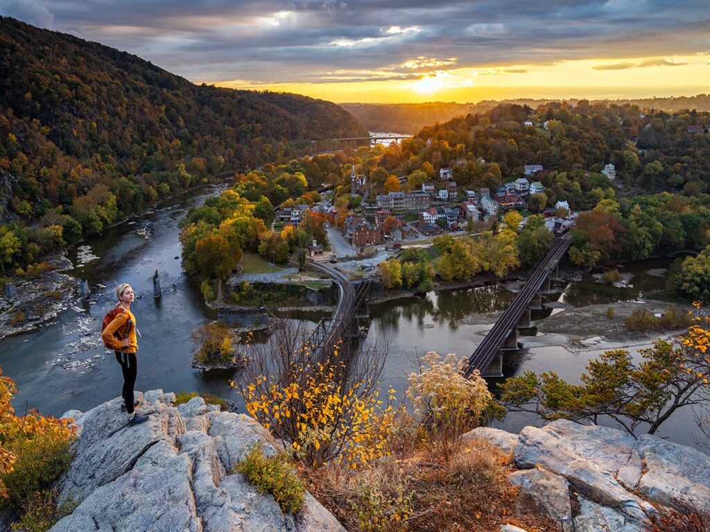 A person stands on a rocky cliff overlooking a scenic river, a small town, and railroad bridges at sunset, with rolling hills and autumn trees in the background.