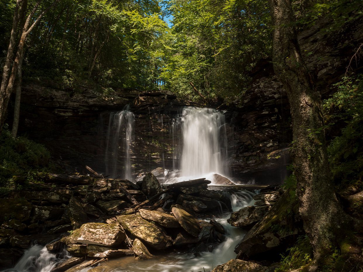 A waterfall cascades over a rocky cliff into a stream surrounded by lush green trees and sunlight filtering through the forest canopy.