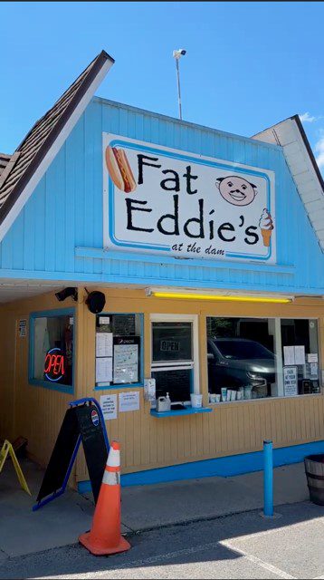 A small blue and tan building with a sign reading Fat Eddies at the dam featuring a hot dog and ice cream cone. There’s a menu, an OPEN sign, and an orange traffic cone outside the window.