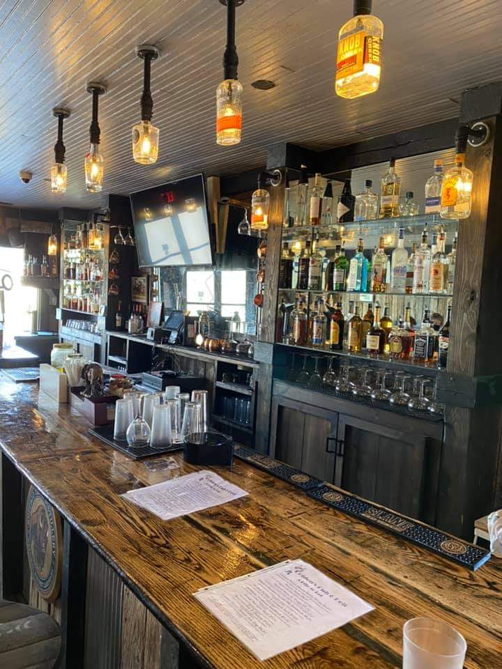 A rustic bar with a wooden counter, glassware, and bottles of liquor displayed on shelves. Warm hanging lights illuminate the area, and a TV is mounted above the bar. A drink menu sits on the counter.