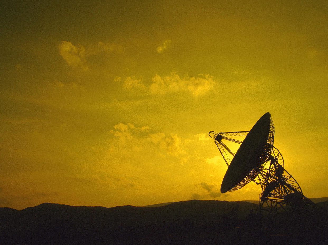 A large radio telescope dish is silhouetted against a yellow sky at sunset, with scattered clouds and distant hills visible in the background.