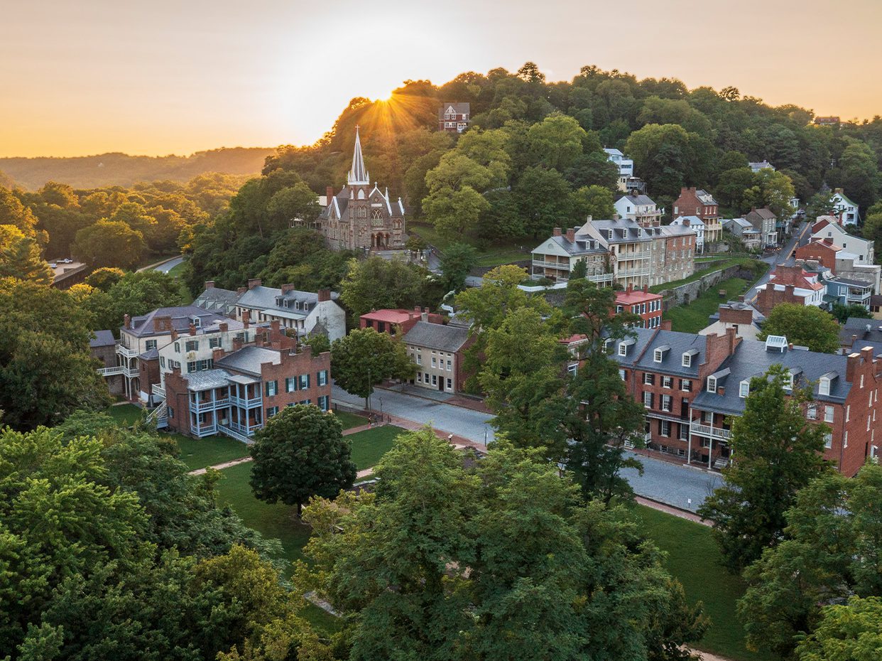 Aerial view of a quaint town with historic brick buildings, a church with a tall steeple, and lush green trees, set against a hillside at sunrise.