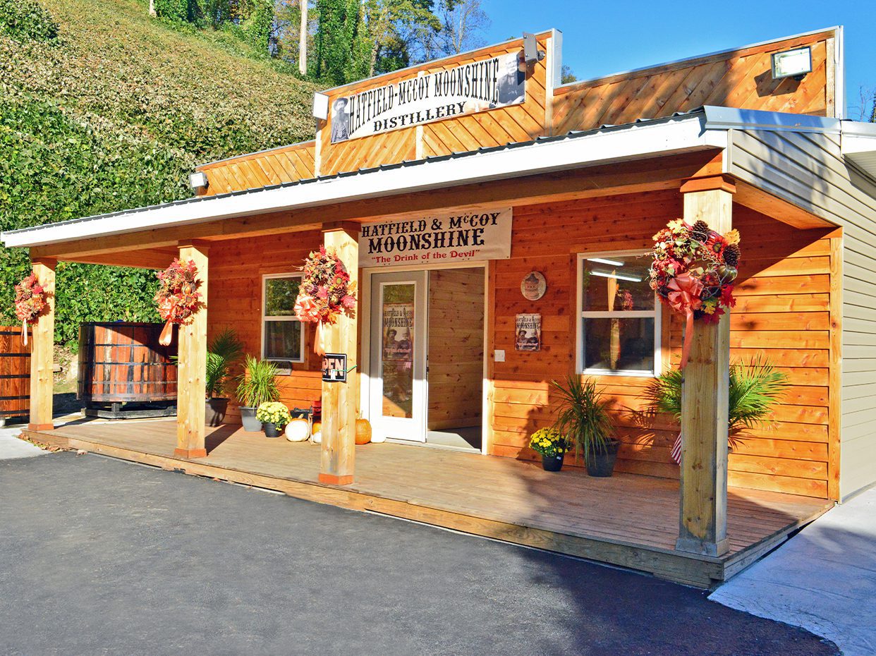 A rustic wooden building with a sign reading Hatfield & McCoy Moonshine Distillery. The entrance is decorated with autumn wreaths, pumpkins, and potted plants, and the building is set against a hill with green foliage.