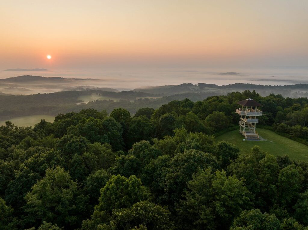 Sunrise over a lush, green forest with rolling hills, mist in the valleys, and a wooden observation tower on a grassy clearing in the foreground.