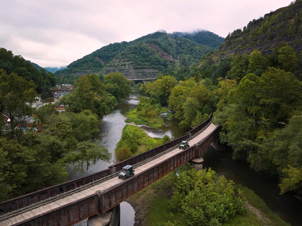 A curved railway bridge crosses a lush, tree-filled river valley with vintage cars driving on it, set against green mountains under a cloudy sky.