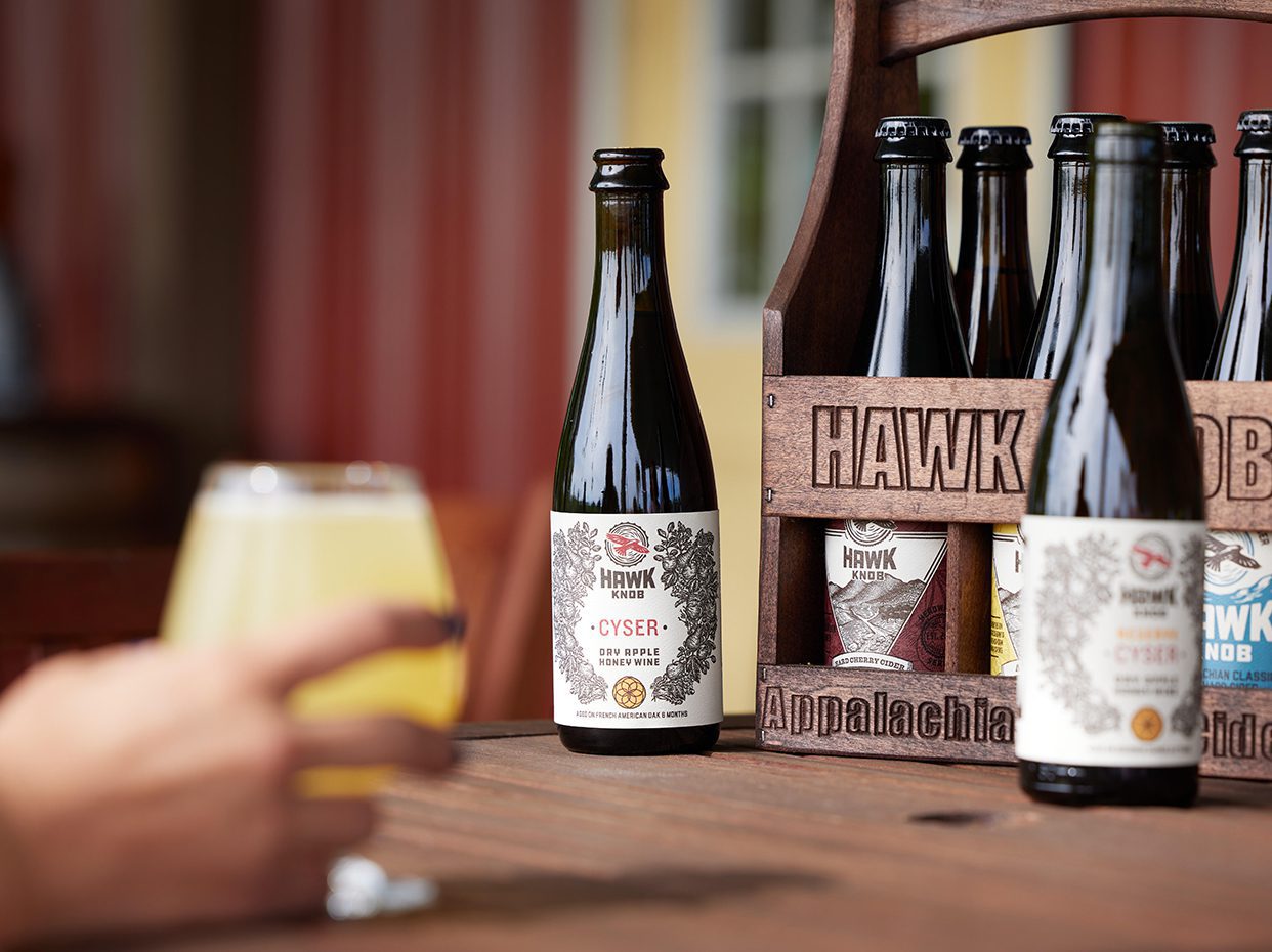 A hand holds a glass of pale yellow beverage next to a bottle labeled “Cyser Sour Apple” and a wooden carrier with more bottles on a wooden table, with striped red and white background.