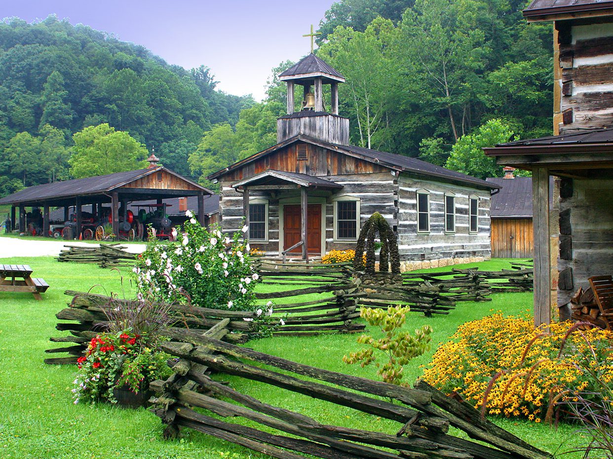 A rustic wooden church with a cross on top sits among gardens and split-rail fences, surrounded by green trees and historic buildings in a rural setting.
