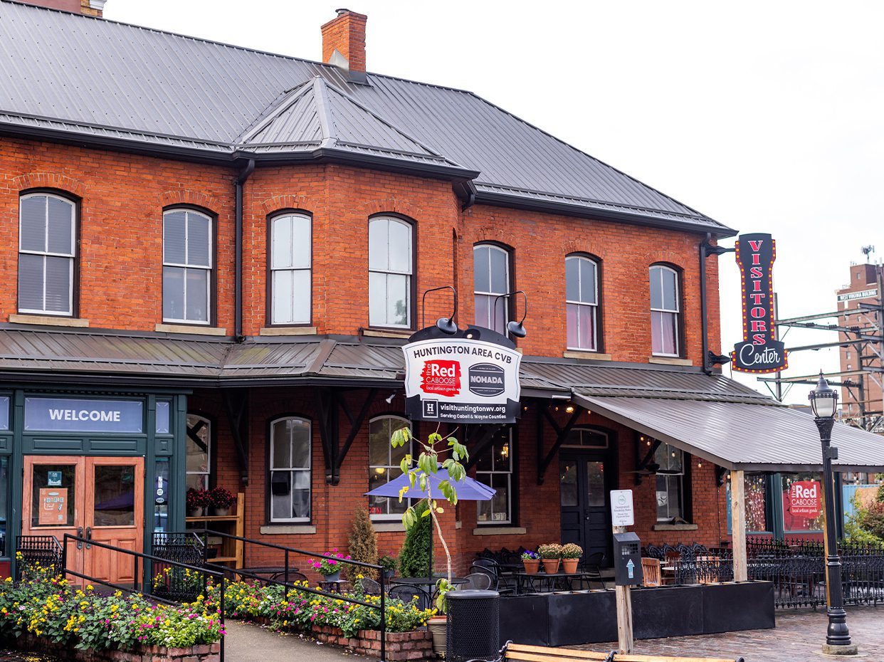 A historic red-brick visitor center building with large windows, a black metal roof, welcome signs, and a hanging Visitors Center sign. There are ramps, potted plants, and a lamppost in front.