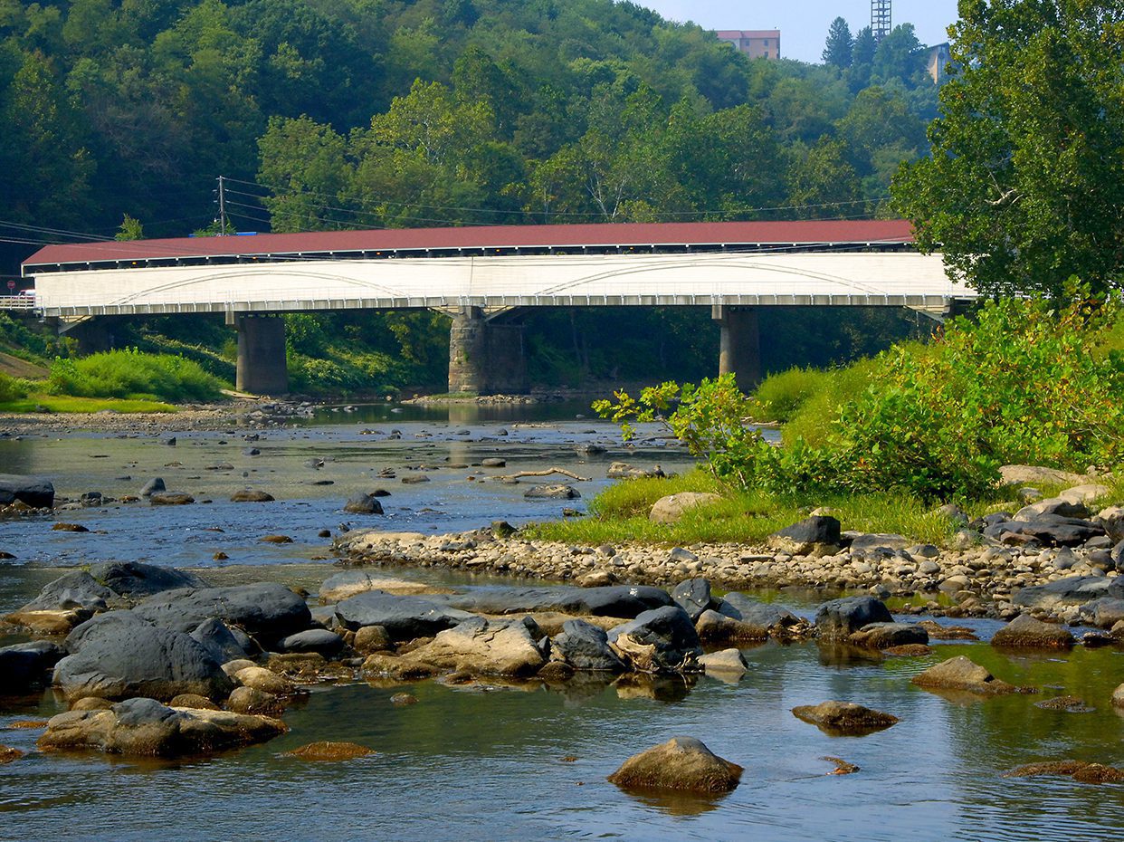A white covered bridge with a red roof spans a rocky river surrounded by green trees and forested hills under a clear sky.