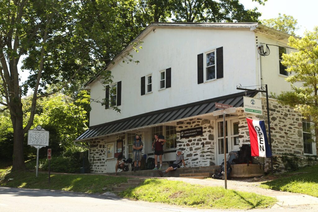 A group of people sits and stands outside a rustic, stone and white wooden building with a metal awning and an OPEN flag. Mature trees shade the building and a historical marker sign stands nearby.