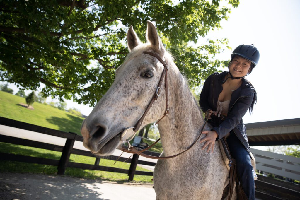A person wearing a helmet smiles while riding a gray horse outdoors near a wooden fence and leafy green trees on a sunny day.