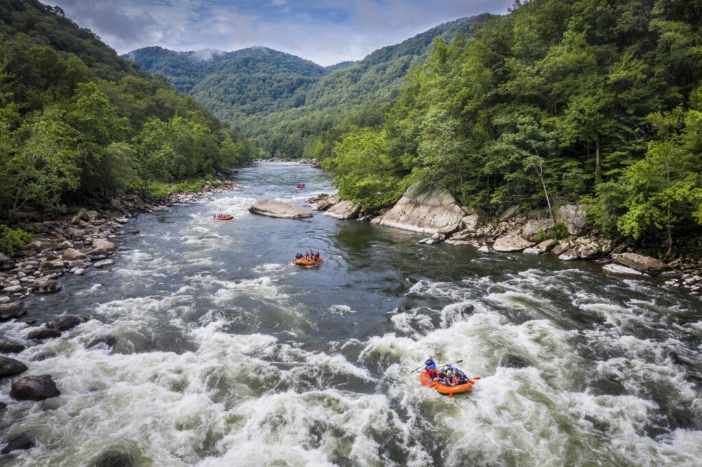 Several groups of people are whitewater rafting on a river surrounded by lush green forest and mountains under a partly cloudy sky.