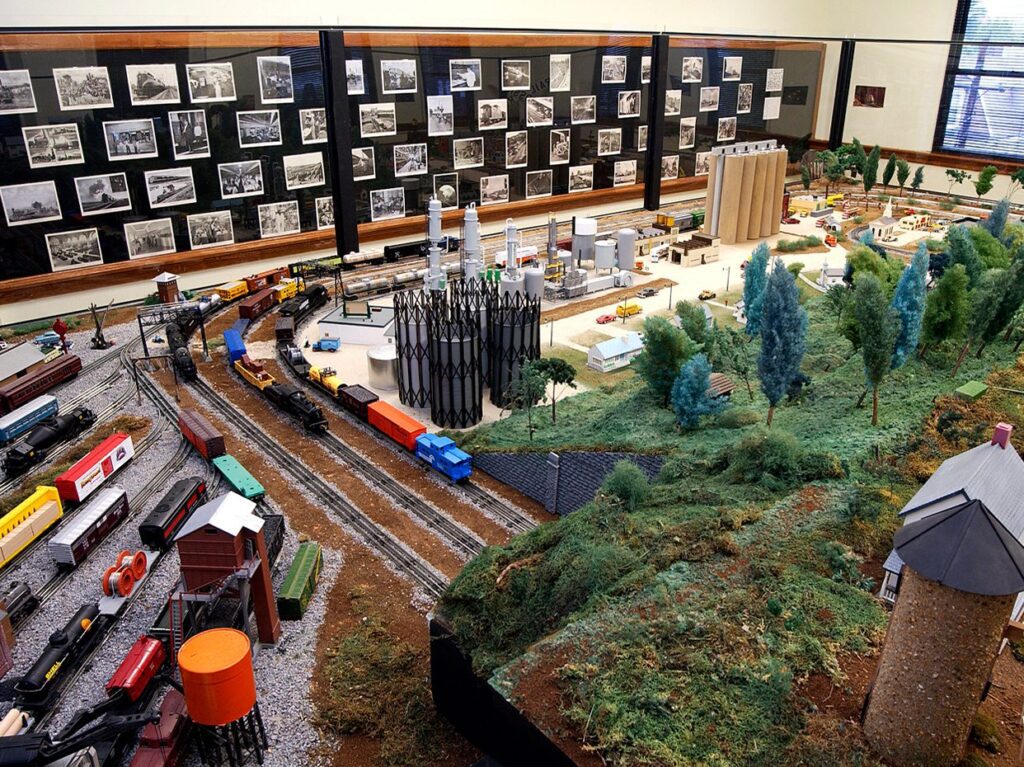 A detailed model train display showcases trains, tracks, buildings, and greenery with a backdrop of framed black-and-white photographs on the wall behind it. Sunlight streams in from a window to the right.