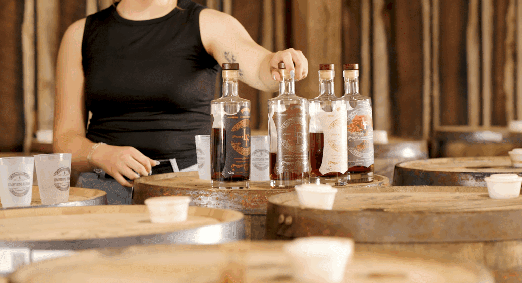 A person in a sleeveless black top stands beside wooden barrels, reaching for one of several whiskey bottles arranged on a barrel, with plastic cups and tasting samples nearby.