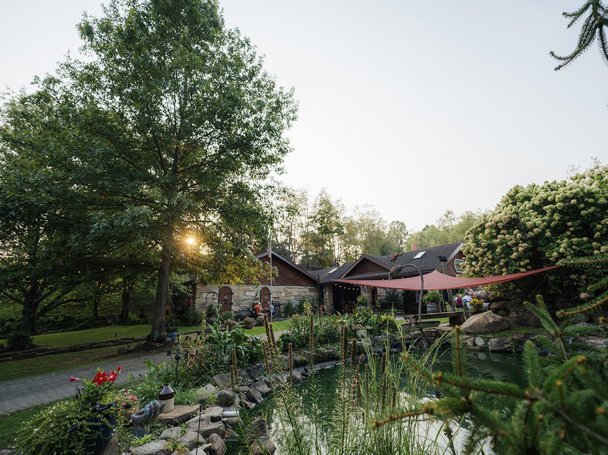 A lush garden with a pond, surrounded by trees and flowering bushes, sits in front of a rustic house at sunset. A red canopy provides shade over a seating area near the pond.