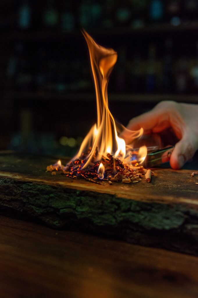 A hand holds metal tongs to light a small pile of wood shavings on fire, with bright orange flames rising from a rustic wooden surface in a dimly lit setting.