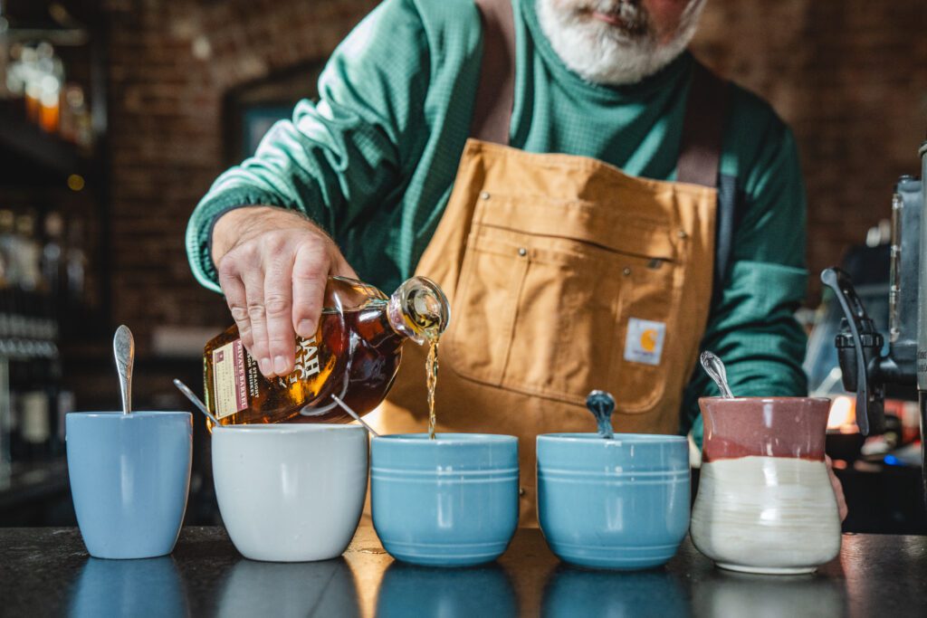 A person in a green sweater and tan apron pours whiskey from a bottle into one of several mugs on a counter, each with a spoon inside, in a cozy, rustic setting.
