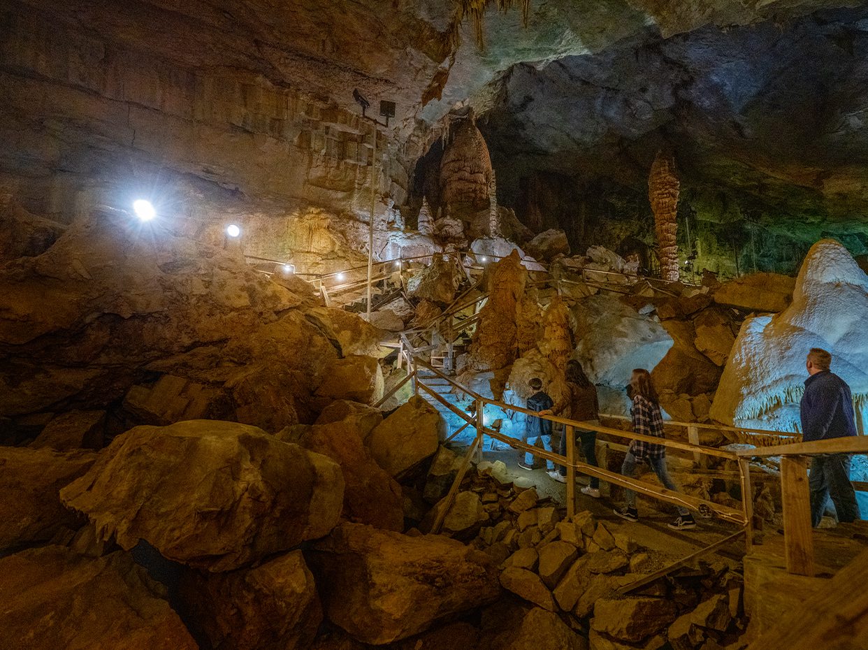 People walk along a fenced path inside a large, rocky cave with stalagmites and uneven ground, illuminated by artificial lights.